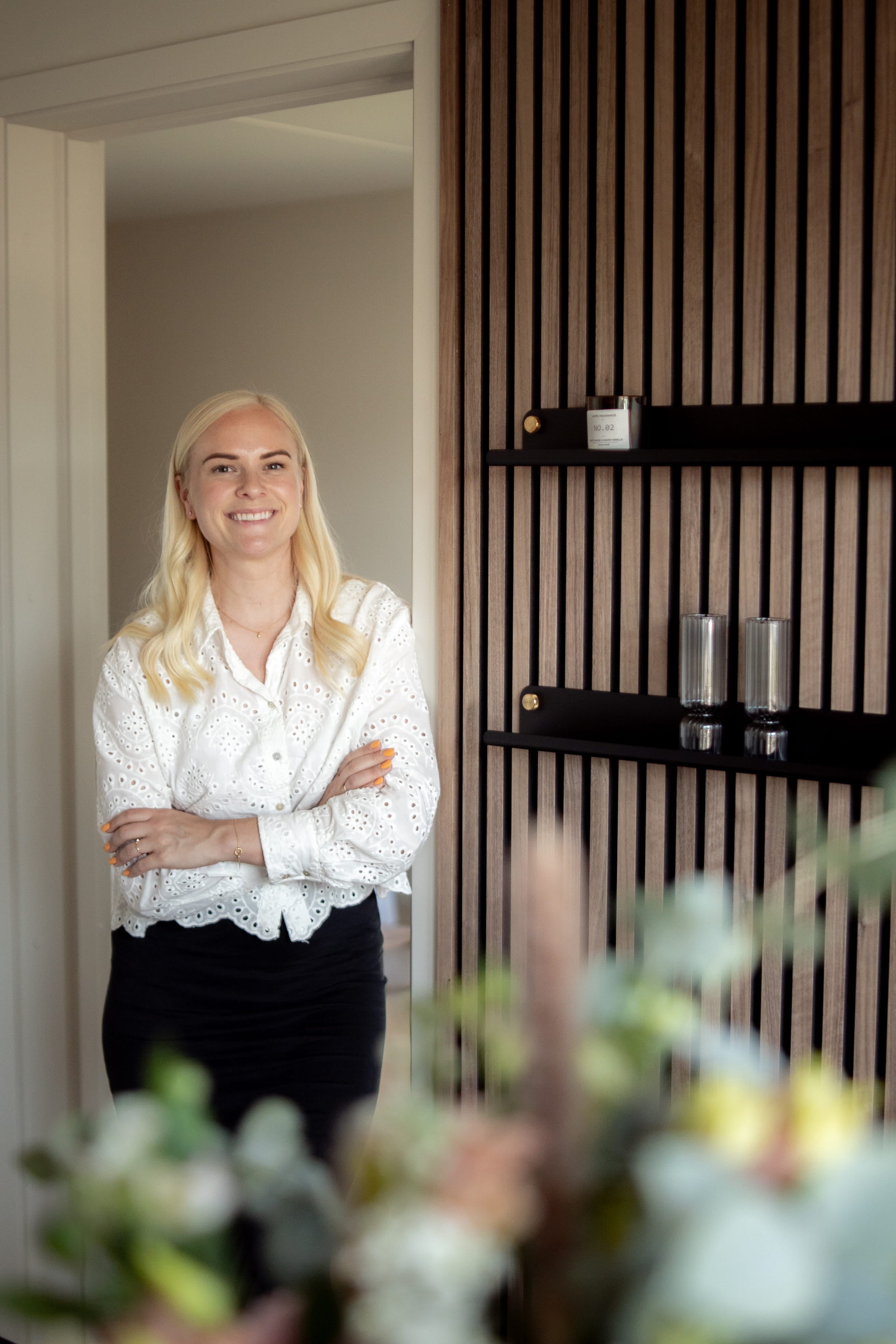 A woman with long blonde hair and a white blouse stands smiling with arms crossed in a modern room featuring Nordic acoustic wood panels and sleek shelves.