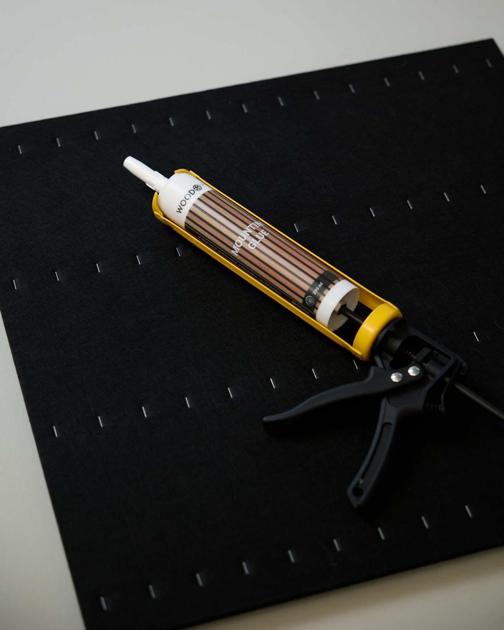 A yellow and black WoodUpp Grout Gun (290 ml) lies on a black mat with a grid of white staples, used for installing acoustic panels.