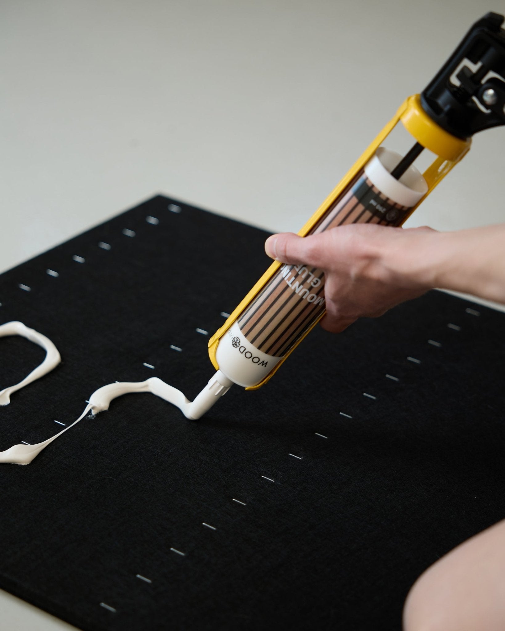 A hand applies white mounting glue in a zigzag pattern with the WoodUpp Grout Gun (290 ml) onto a black acoustic panel surface.