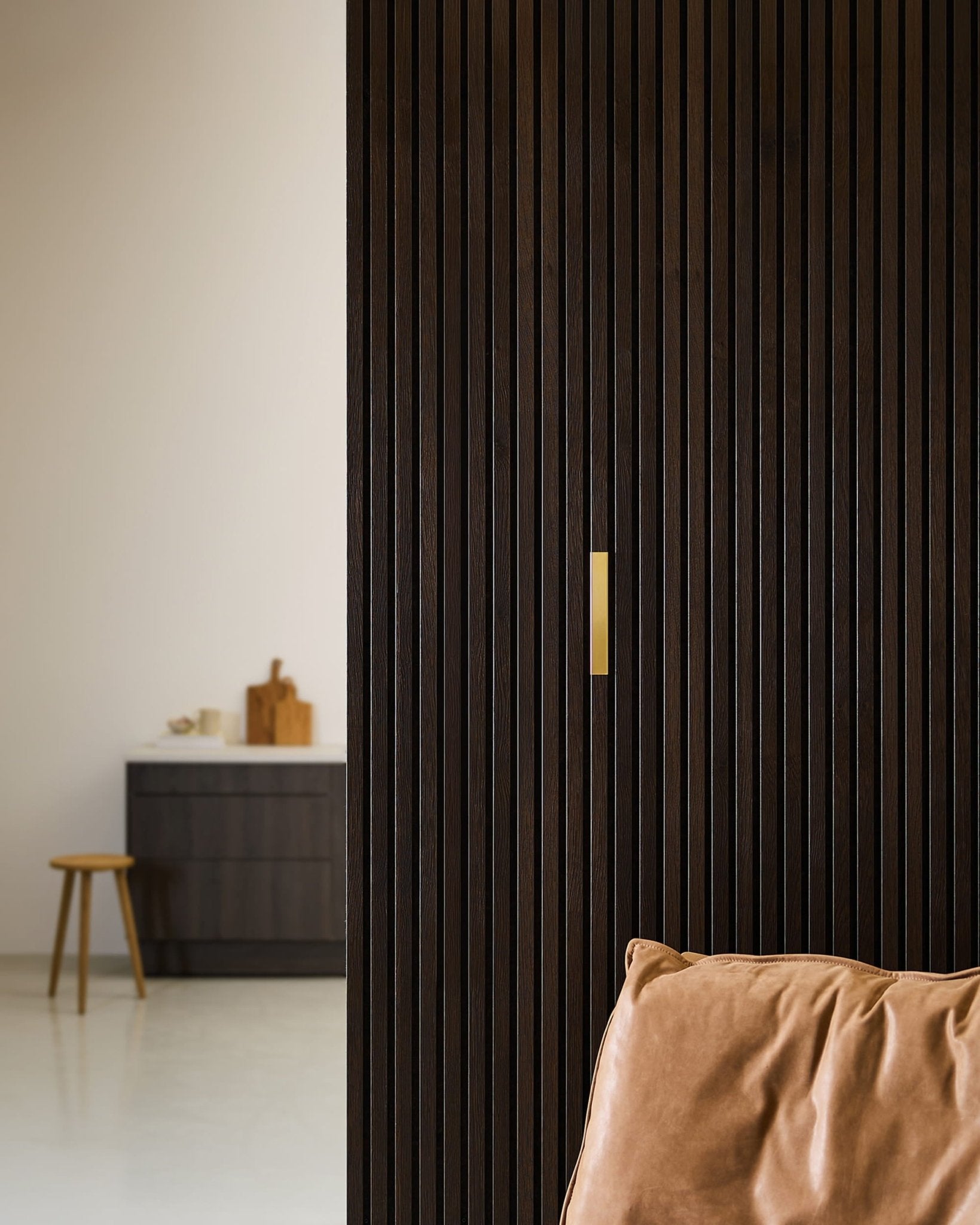 Minimalist room with a WoodUpp Liva | Cover / Brass, 20 cm accessory on a dark wood Akupanel partition, brown leather chair, and black cabinet.
