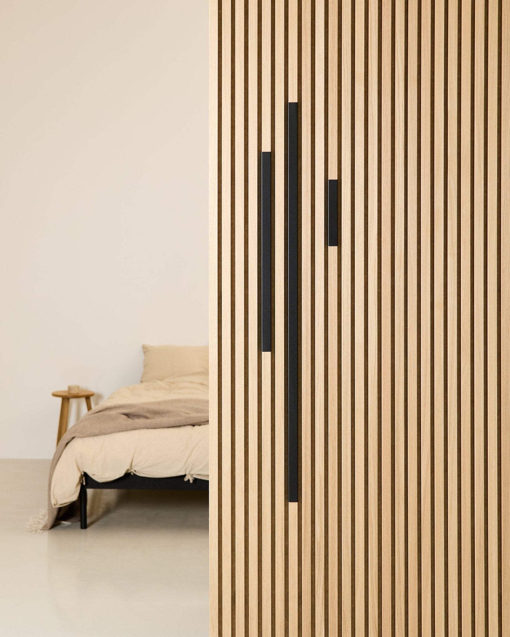 Minimalist bedroom featuring a beige bed and blanket, partially obscured by a 60 cm WoodUpp Akupanel partition in Black Aluminum from the Liva Cover series.