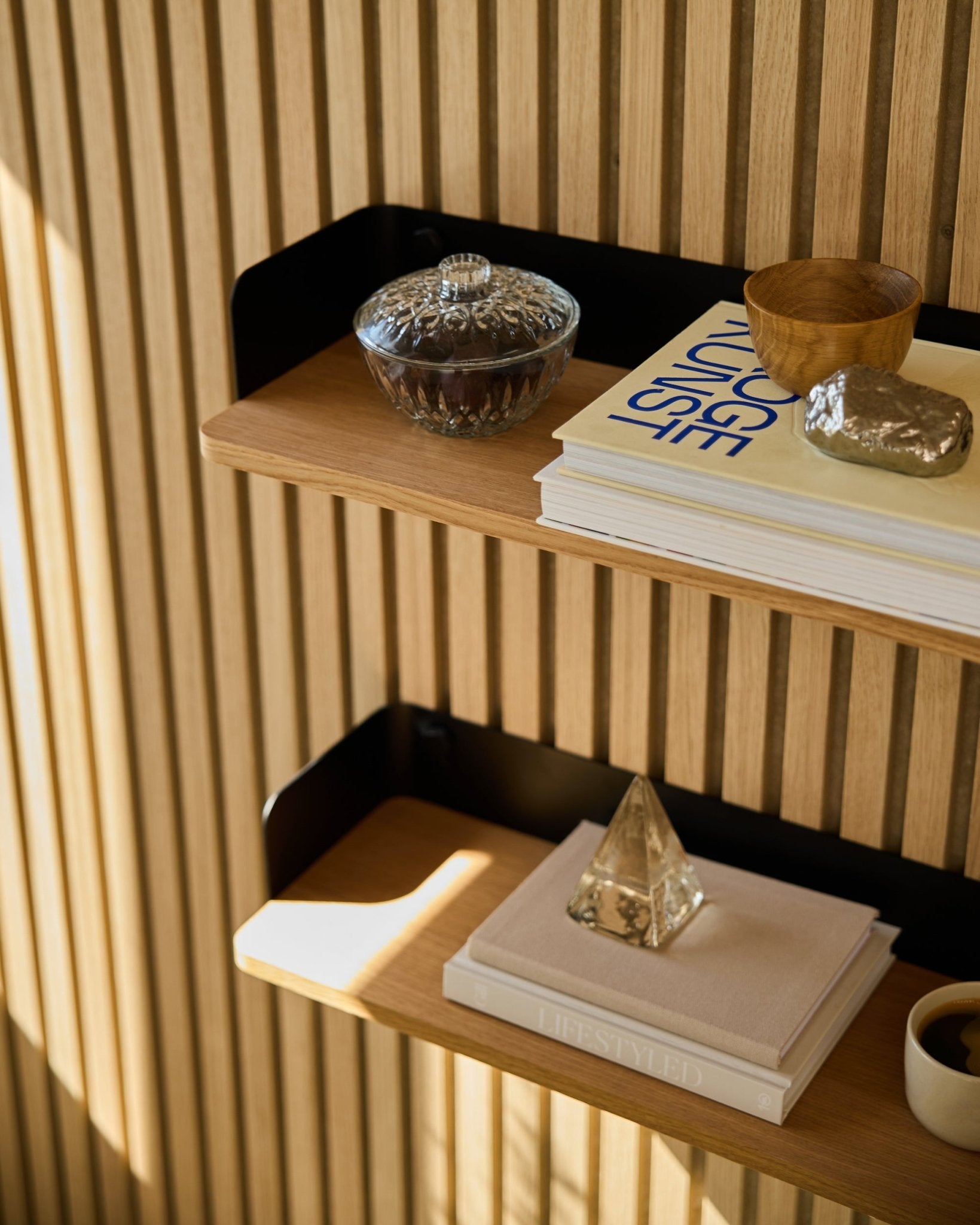 WoodUpp Linn Bookshelf in Natural Oak (55.9 cm) holds a glass jar, cup, gold pyramid, and books against Akupanel acoustic wood panels in sunlight.