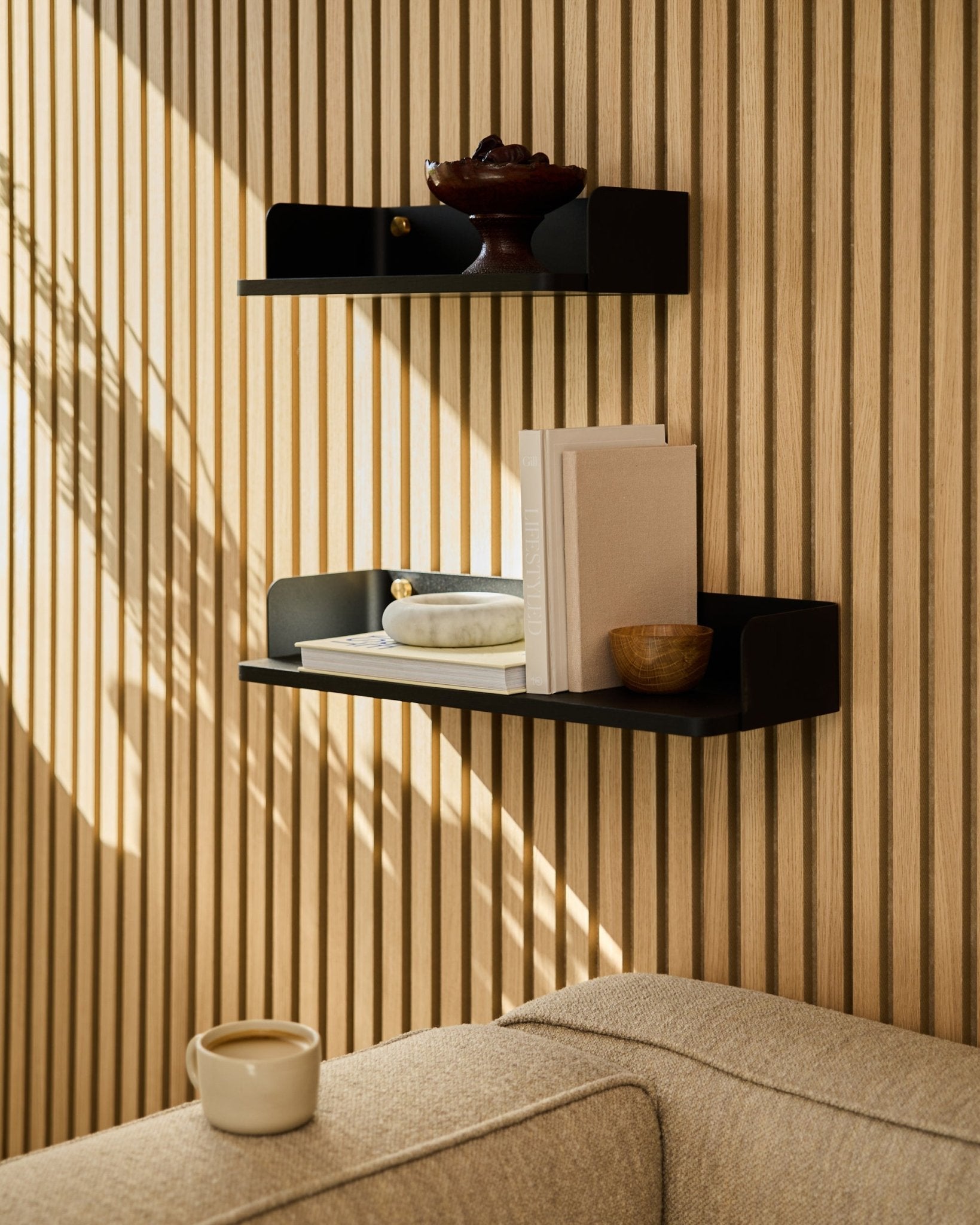 A black oak WoodUpp bookshelf (55.9 cm) displays books, decor, and a bowl on an acoustic wood panel wall; beige sofa and mug in the foreground.