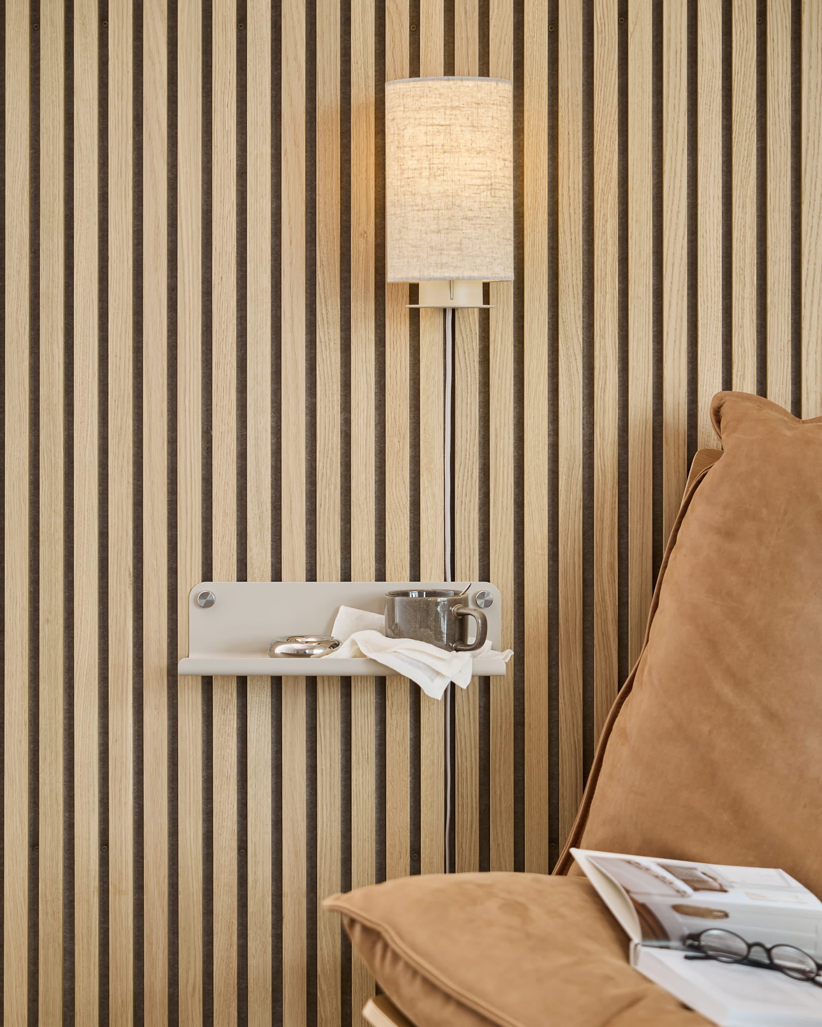 Vertical Akupanel acoustic wood slat wall, beige wall lamp, small shelf holding cup and napkin, tan couch with open magazine.