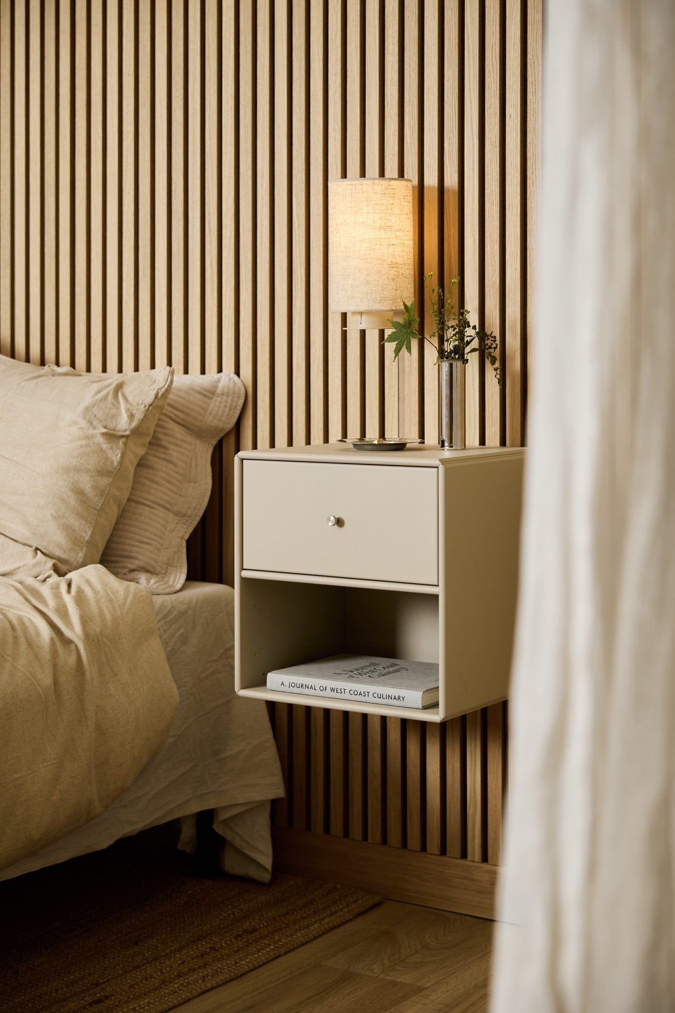 A beige WoodUpp Arka Bookcase Drawer by the bed holds a lamp, books, and vase, set against a WoodUpp Akupanel acoustic wood slatwall panel.