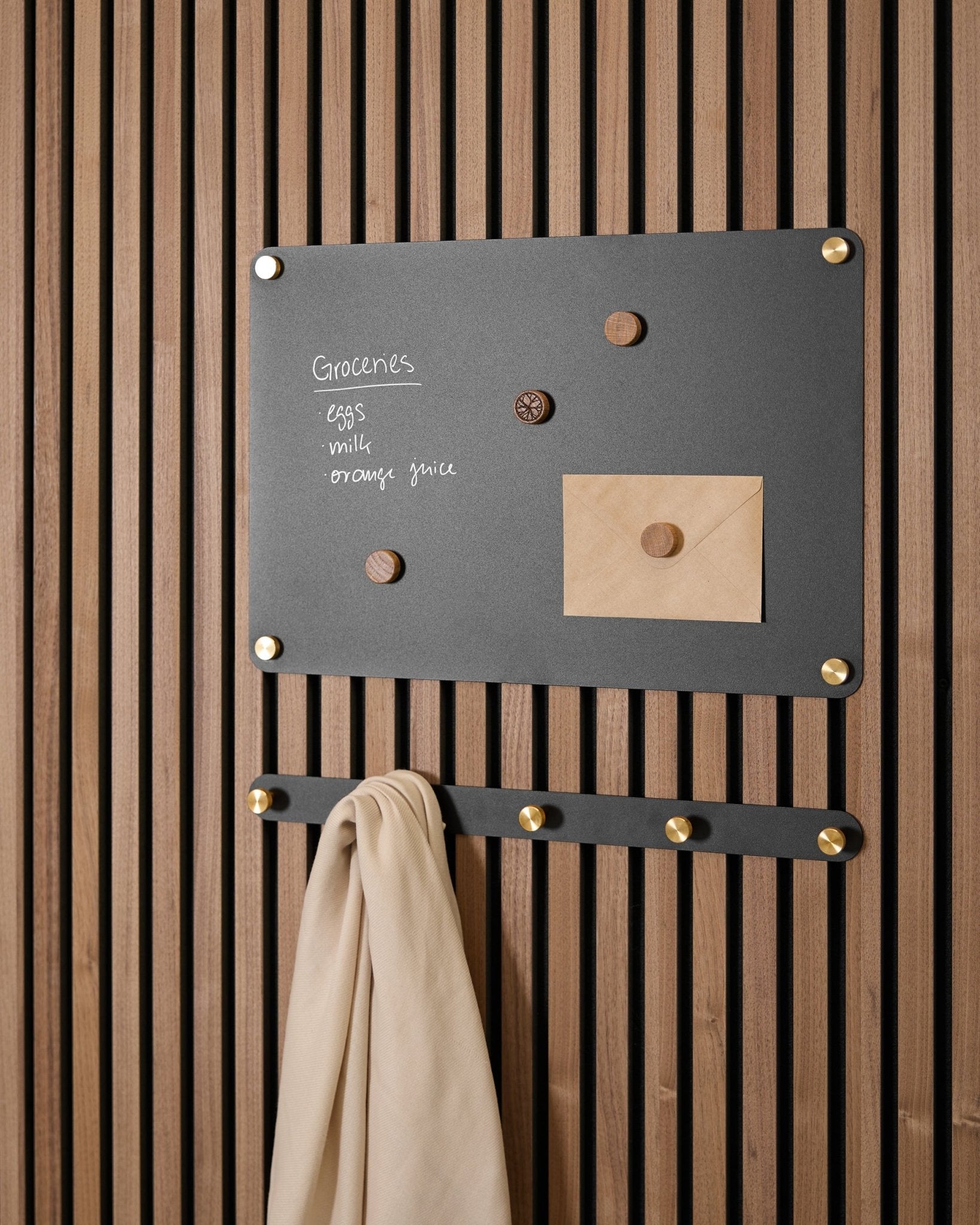 A small black Norr Magnetic Board by WoodUpp holds a grocery list, envelope, magnets, and beige cloth on hooks, mounted on an Akupanel acoustic panel.