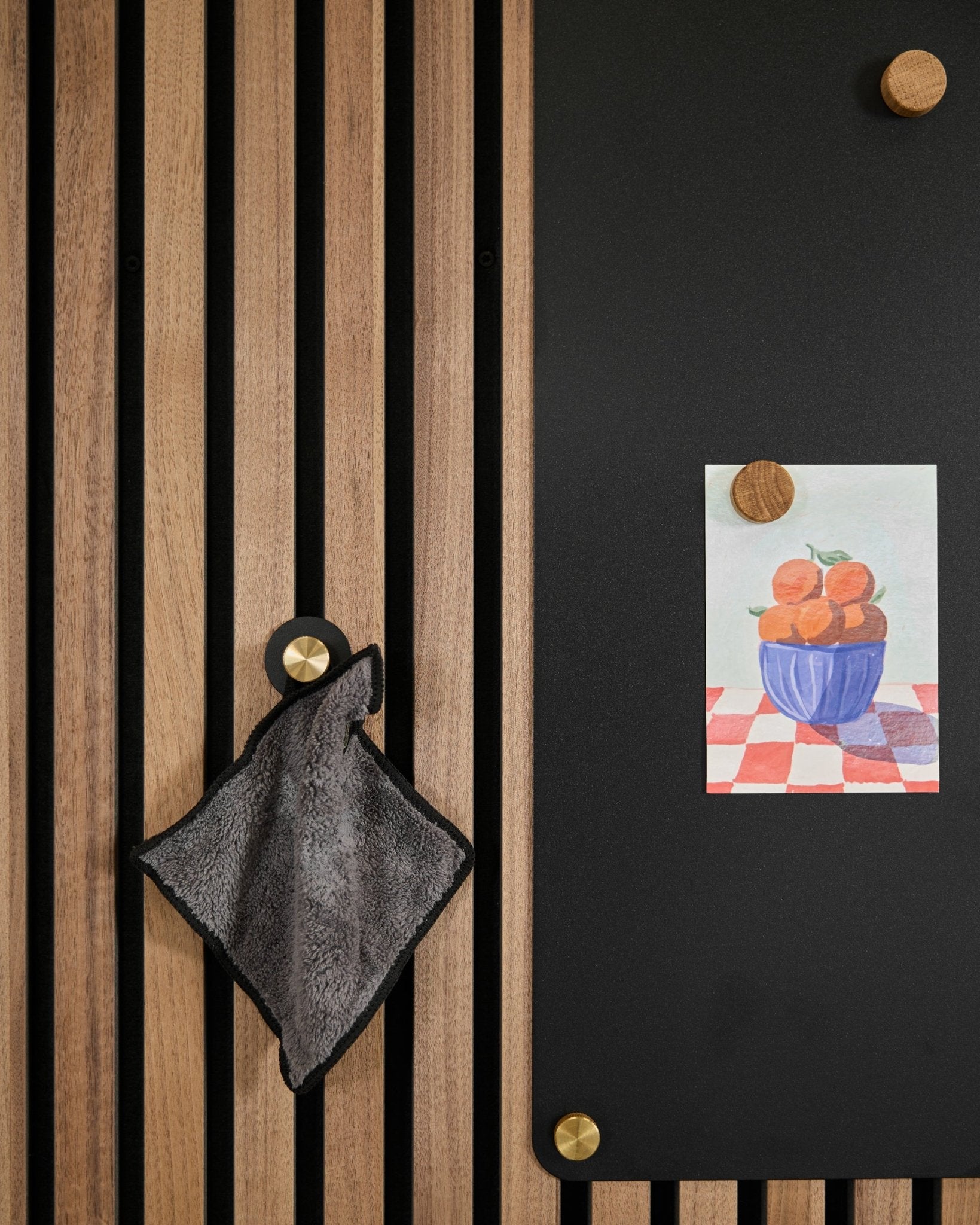 A small towel hangs on a Nordic acoustic slatwall panel beside a black board displaying a drawing of oranges in a bowl, attached with two gold magnets.