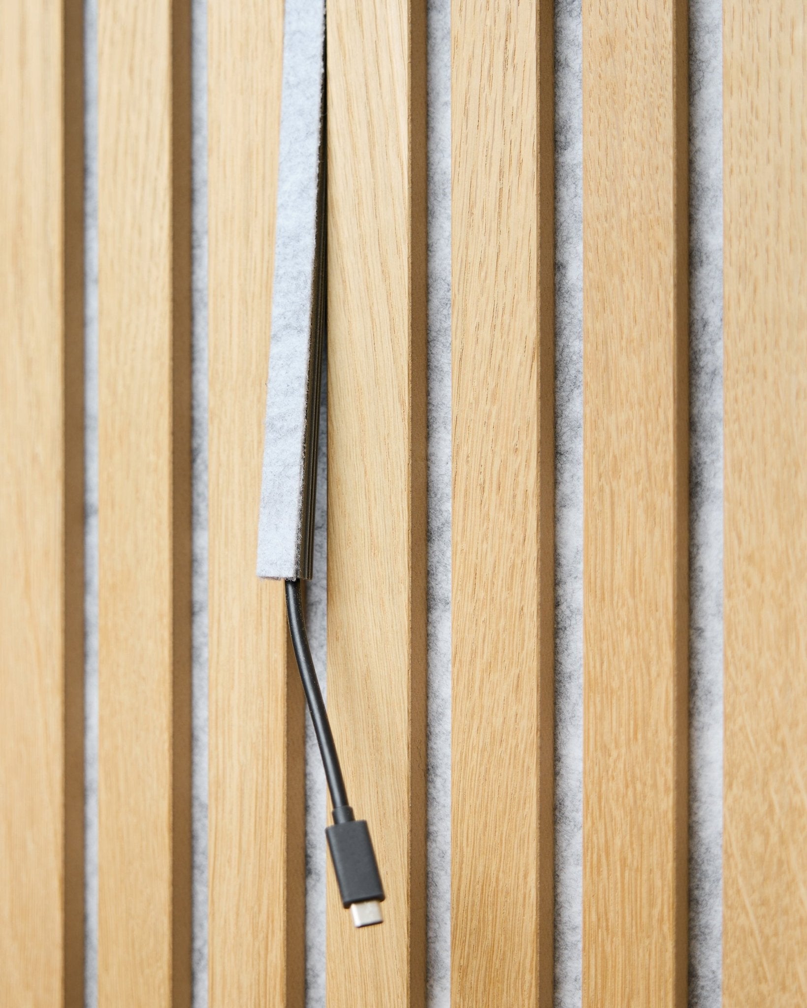 A black USB-C cable runs through a grey WoodUpp Dupps Pop-on Cable Cover between vertical wooden acoustic slats with grey felt backing.