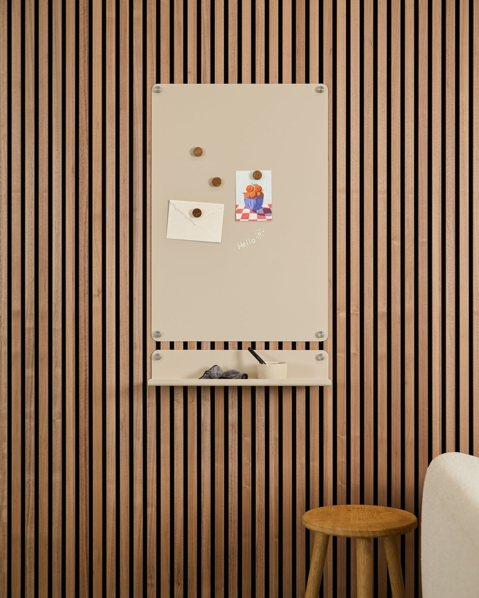 Beige Norr Magnetic Board by WoodUpp mounted above a small shelf on an Akupanel acoustic wood panel wall, with a stool and white sofa nearby.