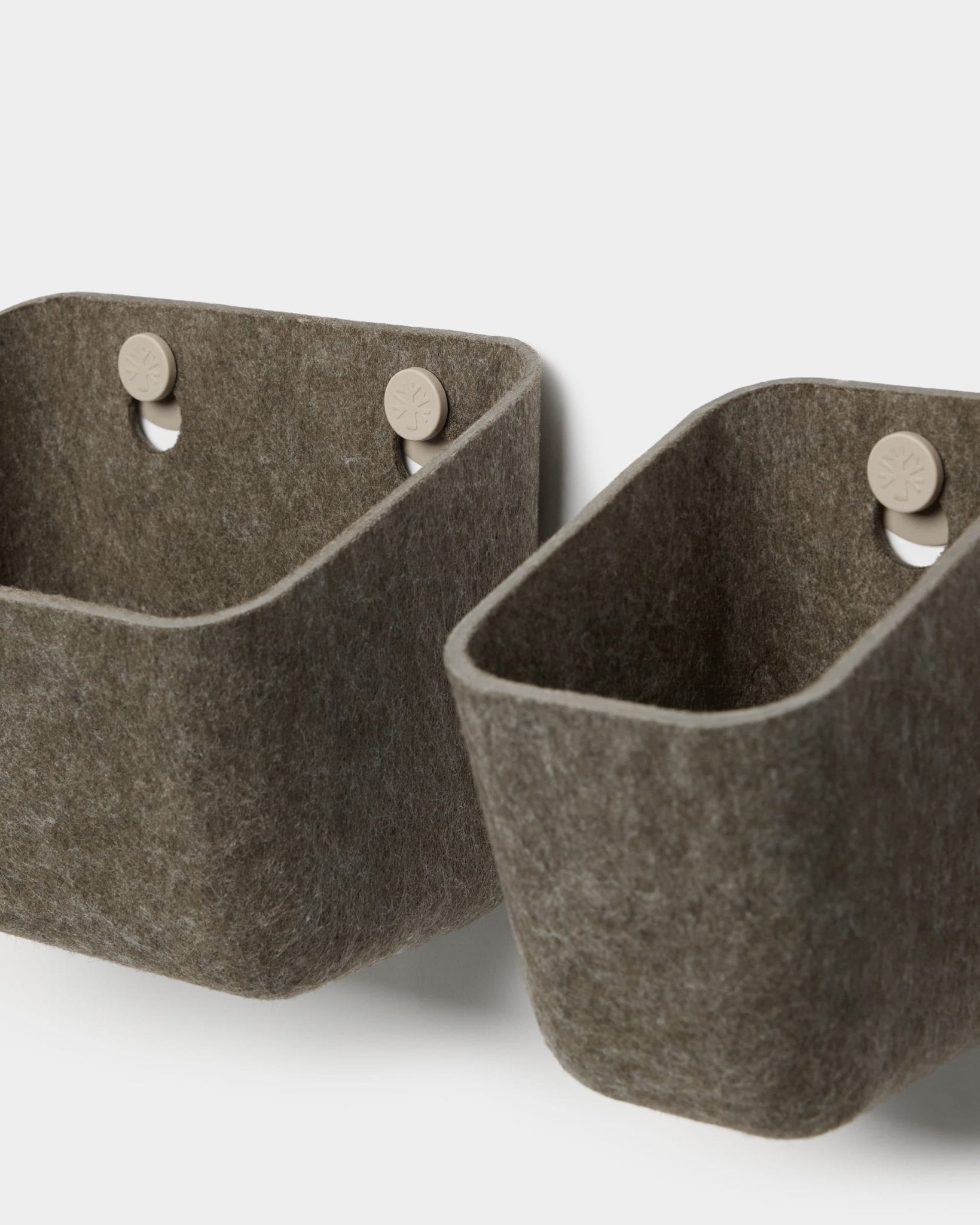 To Luva-filtskabe i mørk, varm grå fra WoodUpp hænger på beige kroge, der er fastgjort til et akustikpanel, og udgør en minimalistisk vægopbevaring med afrundede hjørner.