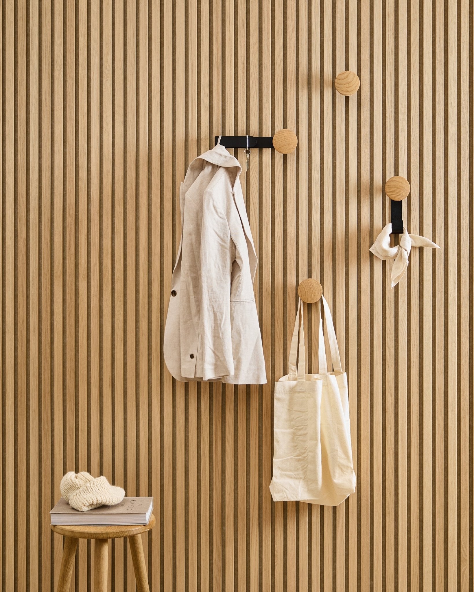 A beige jacket, tote, and scarf hang from WoodUpp’s Linn Horizontal Hanger in Natural Oak; a nearby stool supports a book and a knitted item.