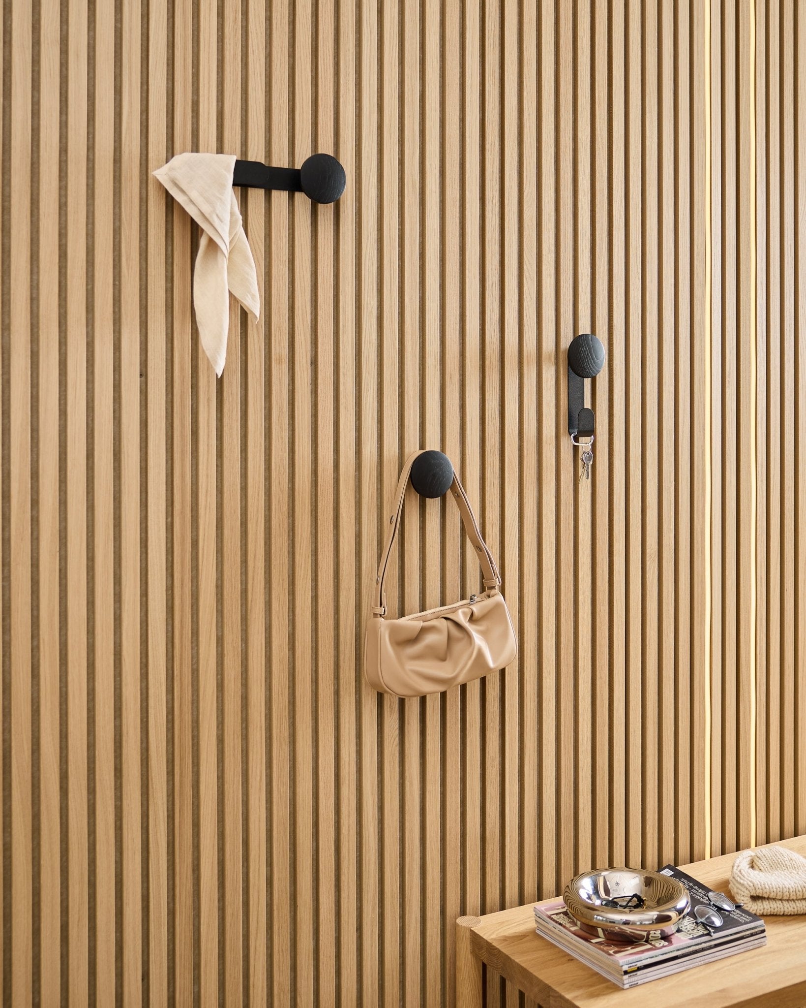 WoodUpp Linn Single Hanger in Black Oak holds a scarf, beige bag, and keys on a black Akupanel acoustic wood panel; bench with books and metal dish nearby.