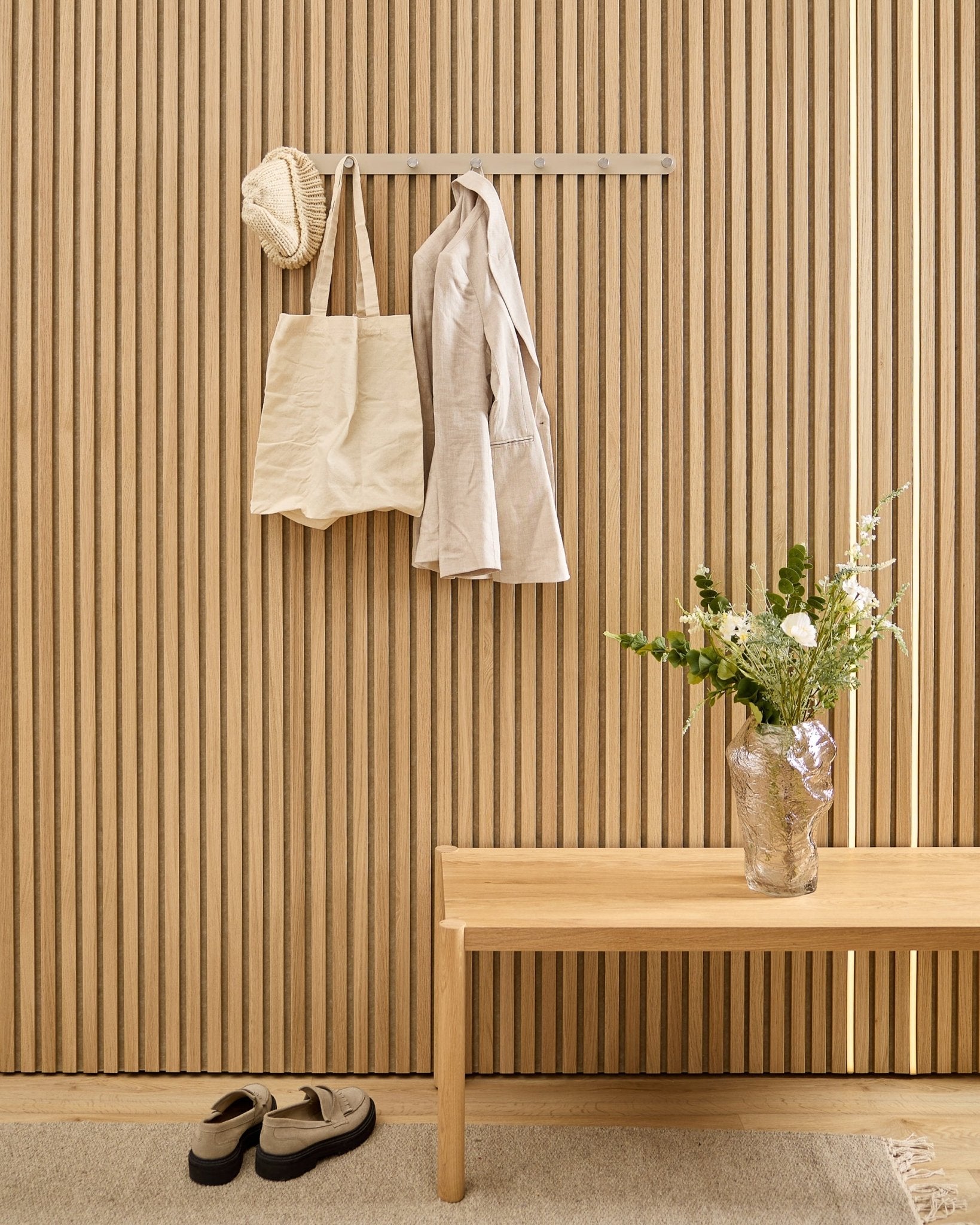 A beige bag, hat, and shirt hang on the WoodUpp Norr Coat Hook Rack above shoes, with a bench and vase beside the striped acoustic slatwall panel.