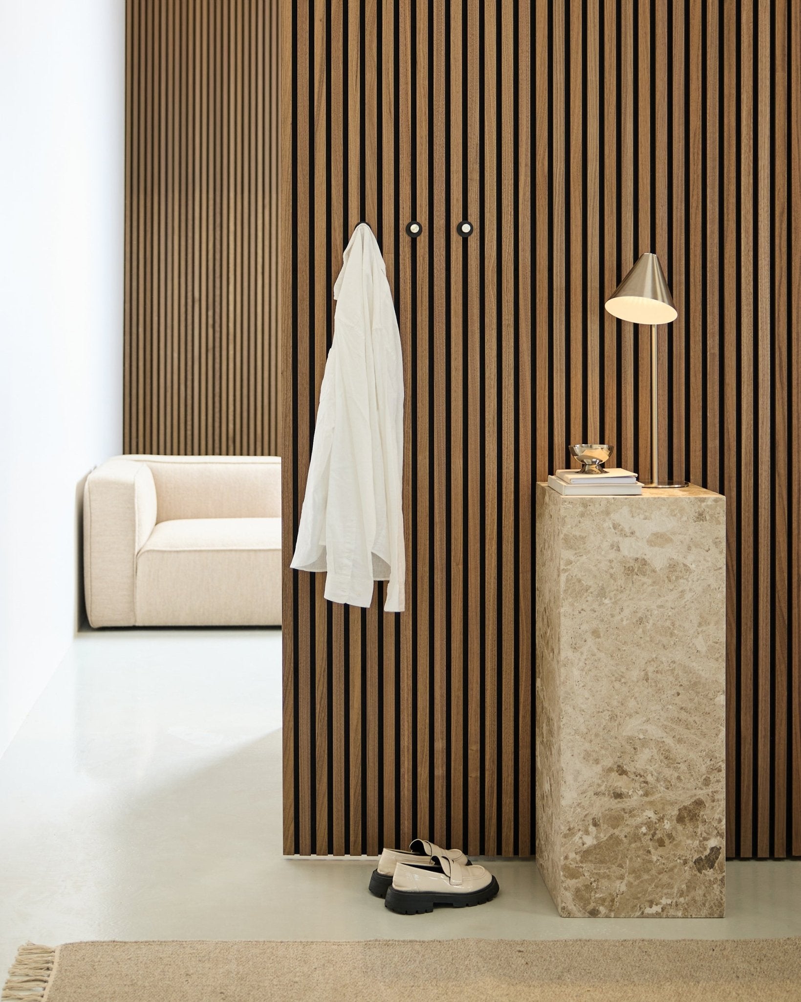Minimalist room with a Nordic Akupanel wood slat acoustic wall, white shirt on wall hooks, stone pedestal holding lamp and books, beige sofa, and white shoes on floor.