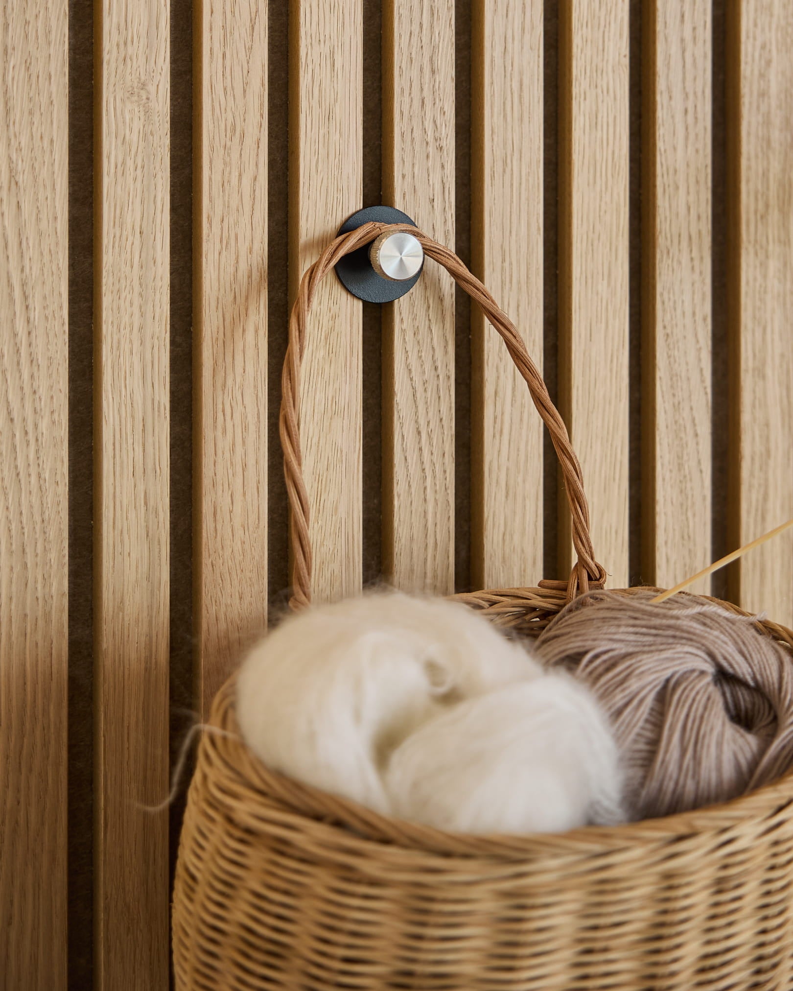 A white and beige yarn wicker basket hangs on a Norr Coat Hook / X1, Black/Stainless Steel by WoodUpp mounted on an Akupanel acoustic slatwall panel.