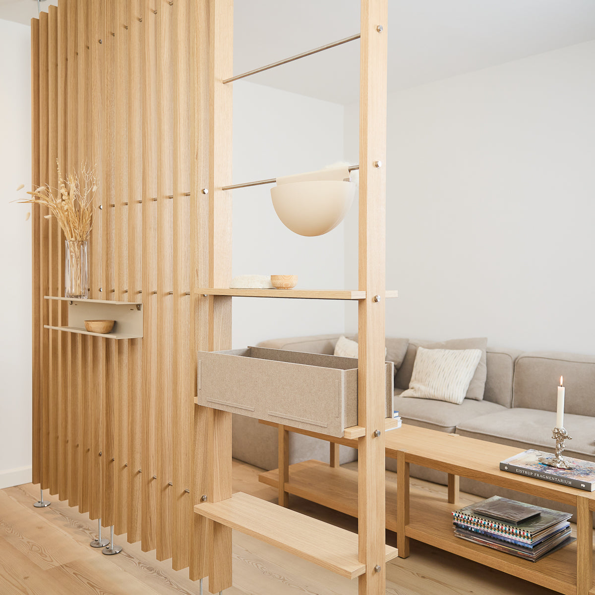 Modern living room featuring a light wood EMBRACE slatted room divider, beige sofa, wooden coffee table, books, candle, and dried flowers in a vase.
