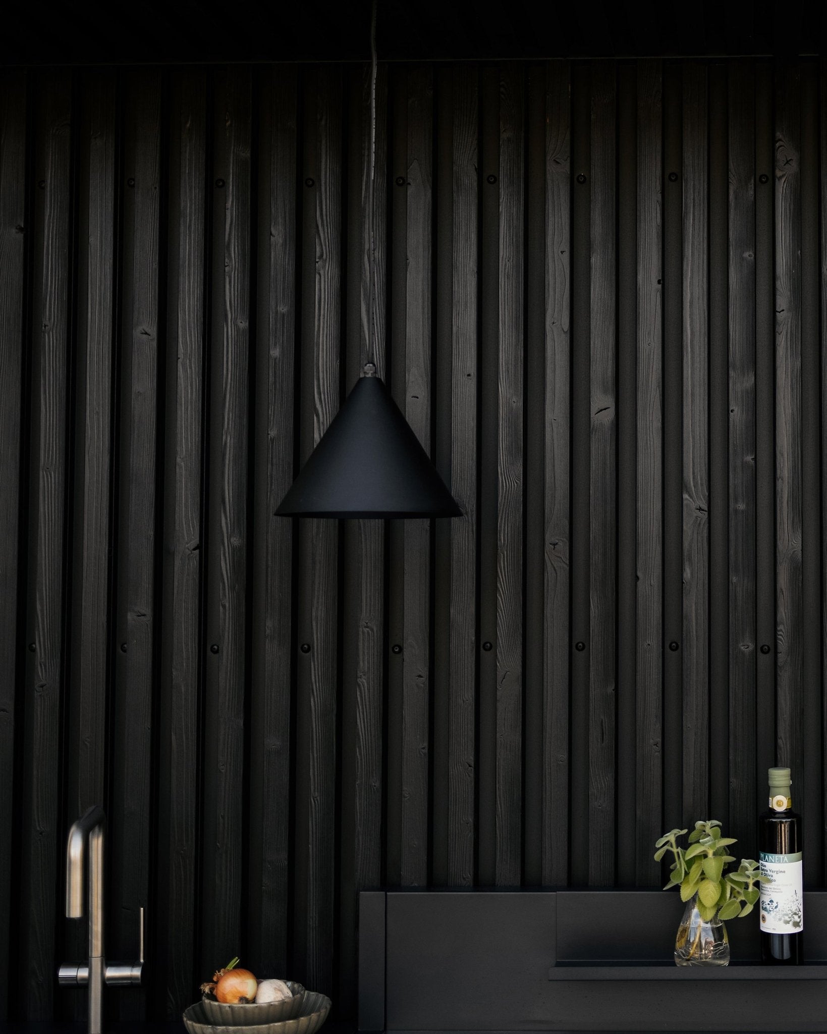 Modern kitchen with matte black pendant lamp, textured black Akupanel acoustic wood wall, and countertop holding vegetables, fresh basil, and olive oil.