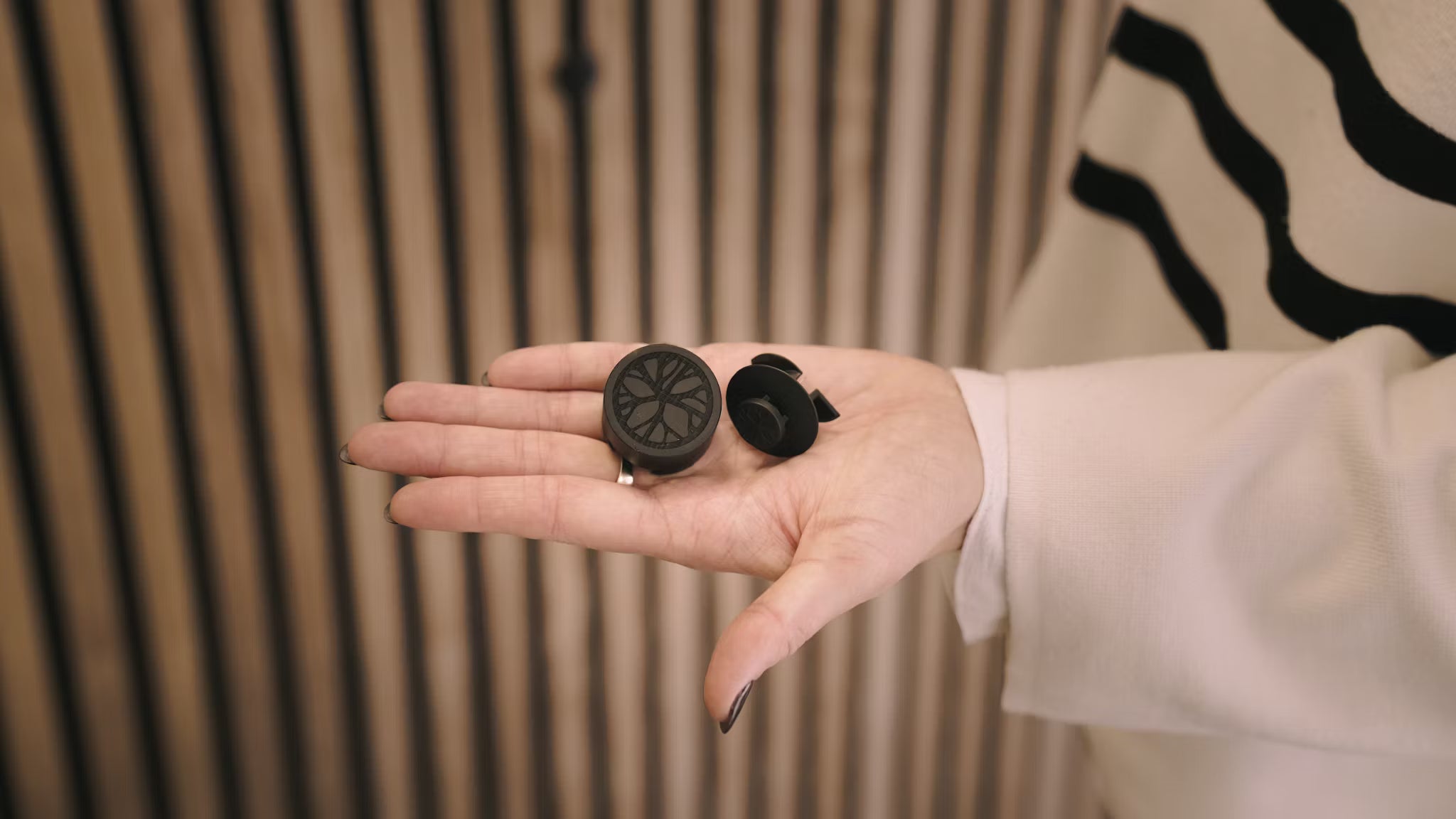 A person in a white sleeve holds two small black gadgets in their open palm with vertical acoustic wood panel slats from WoodUpp’s Akupanel collection behind.