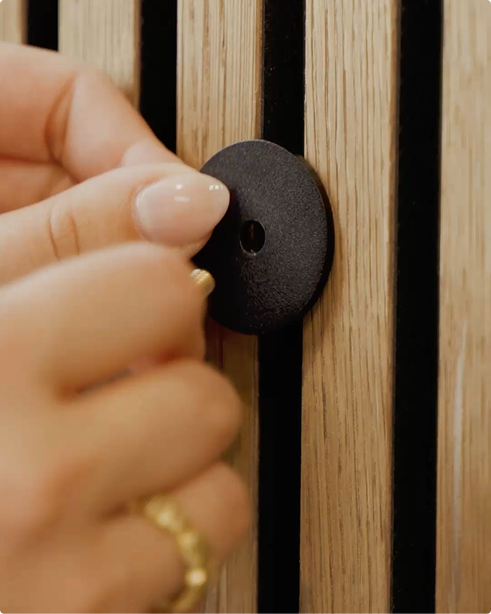 A hand attaches a round black hook to a vertical acoustic slatwall panel, part of the WoodUpp Akupanel collection.