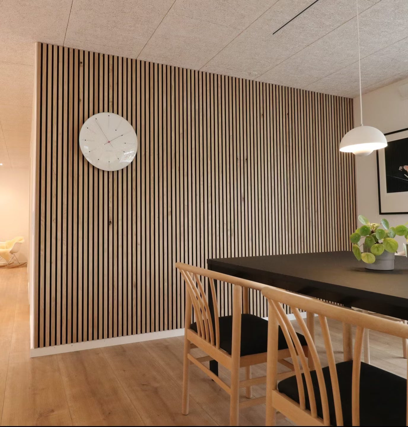 Modern dining room with a Nordic Akupanel acoustic wood slat accent wall, black table, wooden chairs with black seats, and a potted plant centerpiece.