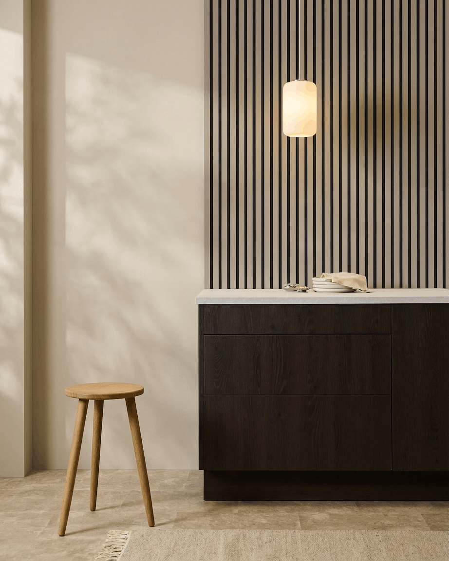 Minimalist kitchen featuring WoodUpp Akupanel (Stone, Black Felt) acoustic panels on the wall, wooden stool, dark cabinets, and pendant light in neutral tones.
