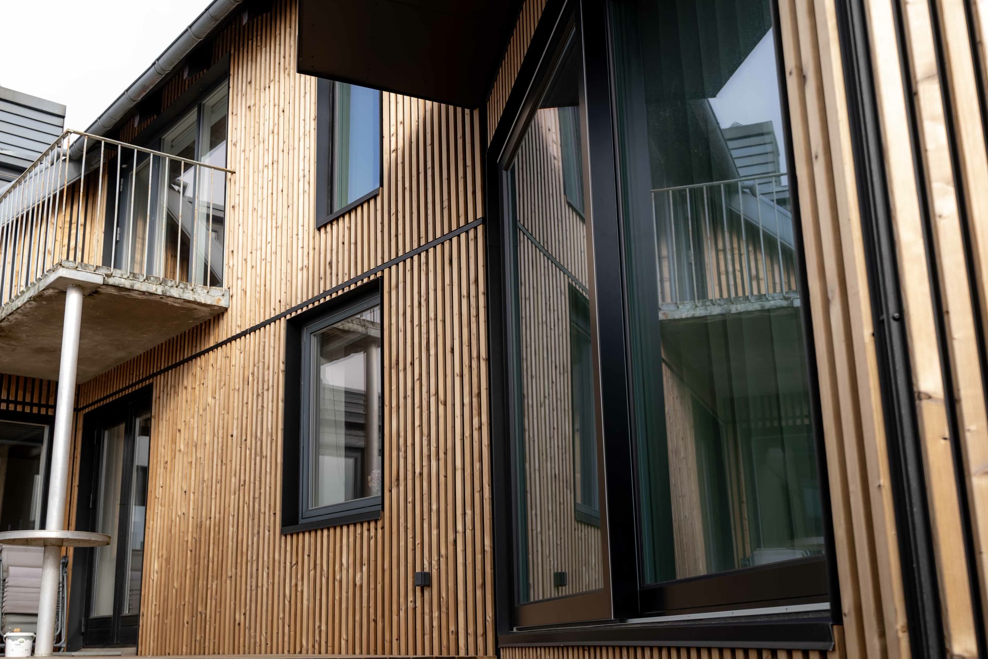 Angled view of a house facade clad in vertical oiled Aluwood slats with a balcony and black window frames