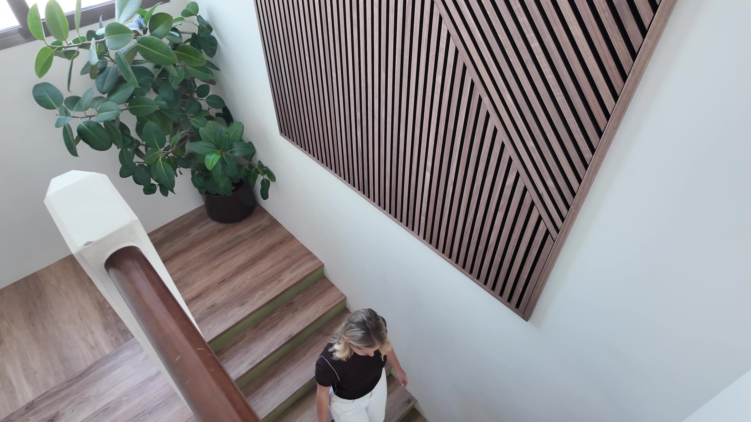 Looking down a stairwell from above, a woman walking down the stairs past a large framed Akupanel slat artwork with slats at several angles on the wall