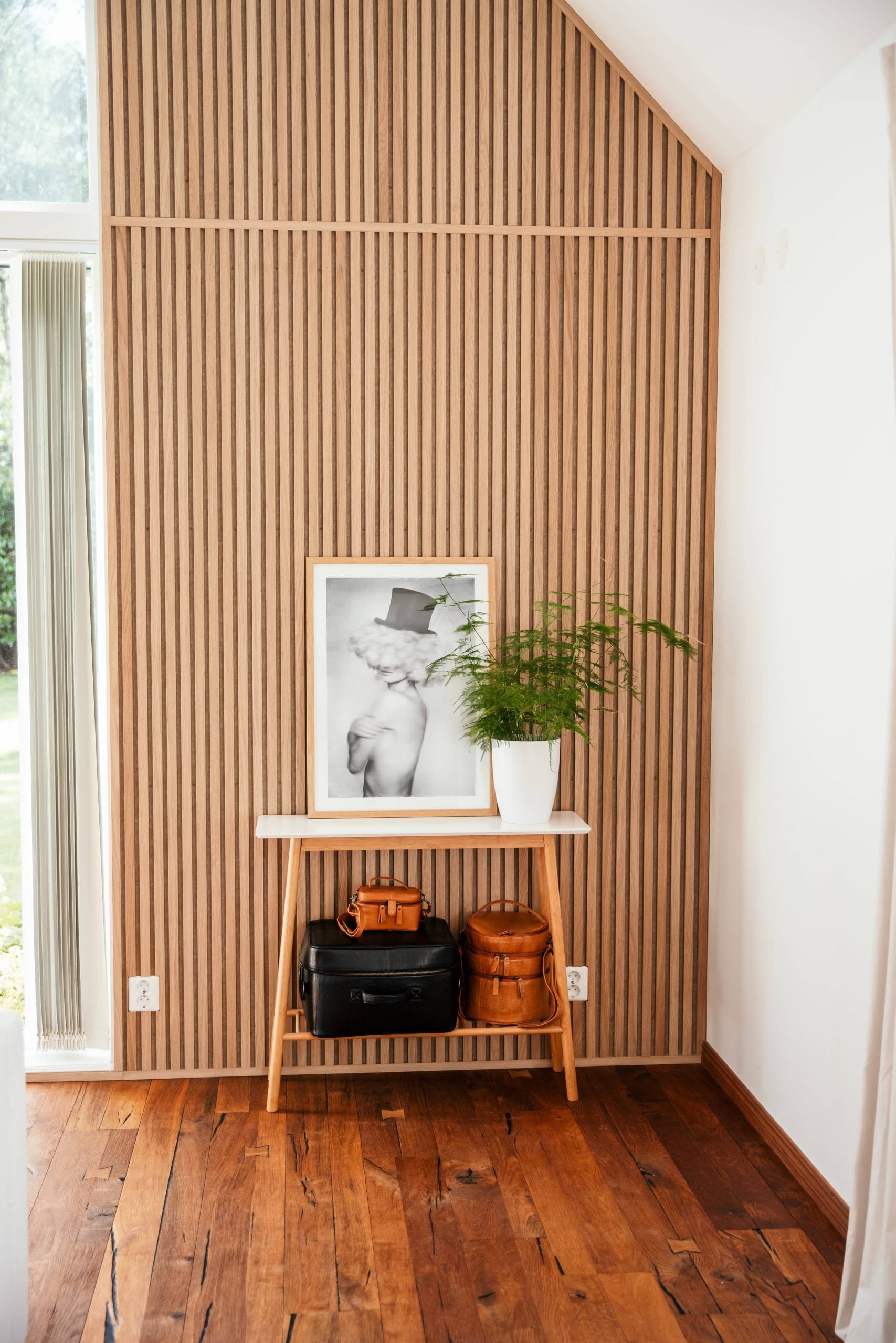 A small wooden table with bags, a framed photo, and a potted plant stands against a Nordic acoustic slatwall panel in a bright room with wooden floors.