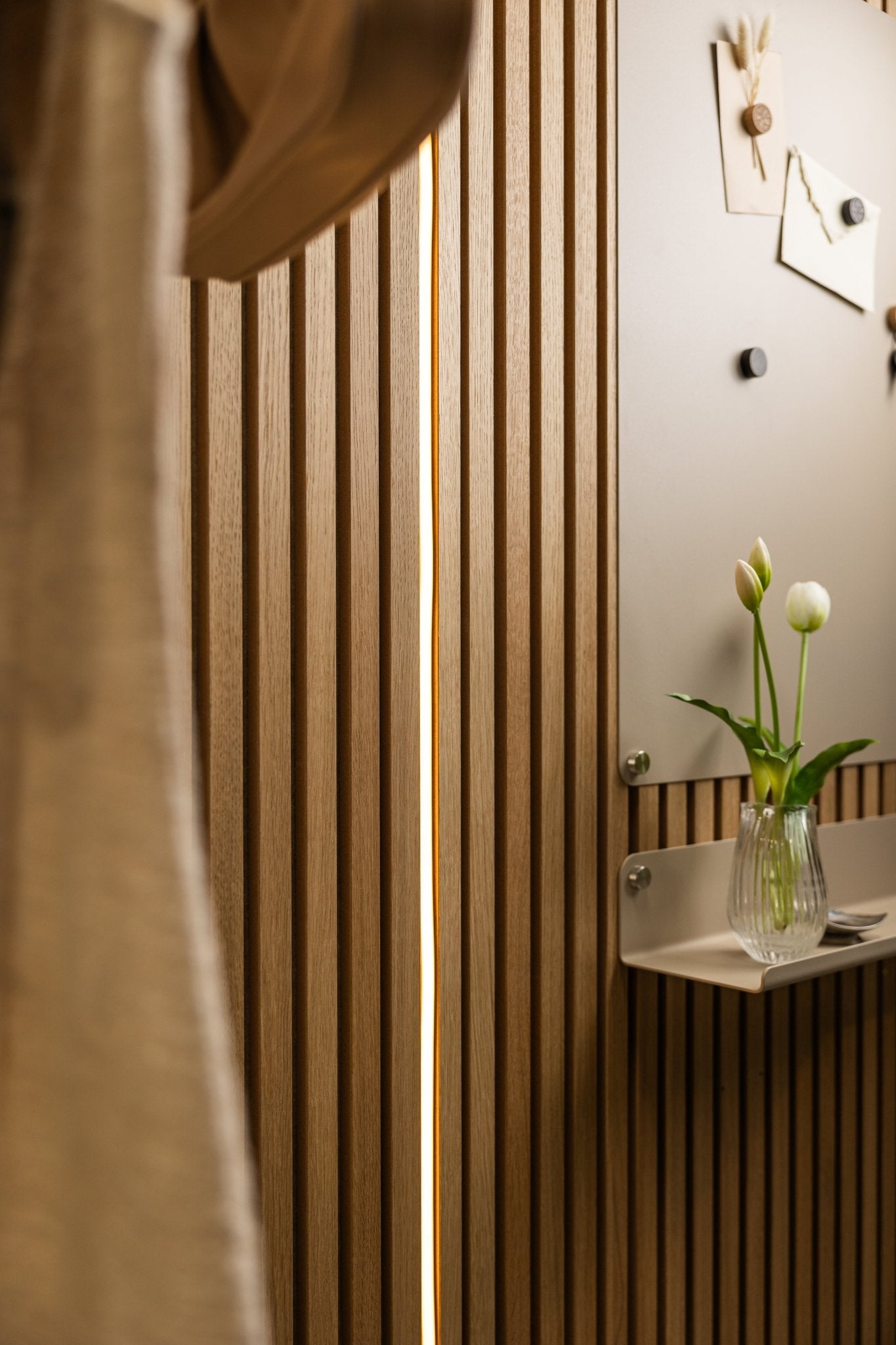 WoodUpp Akupanel acoustic wood slat panel with beige Lumi LED striplights (240 cm), a small shelf holding tulips, and a bulletin board above.