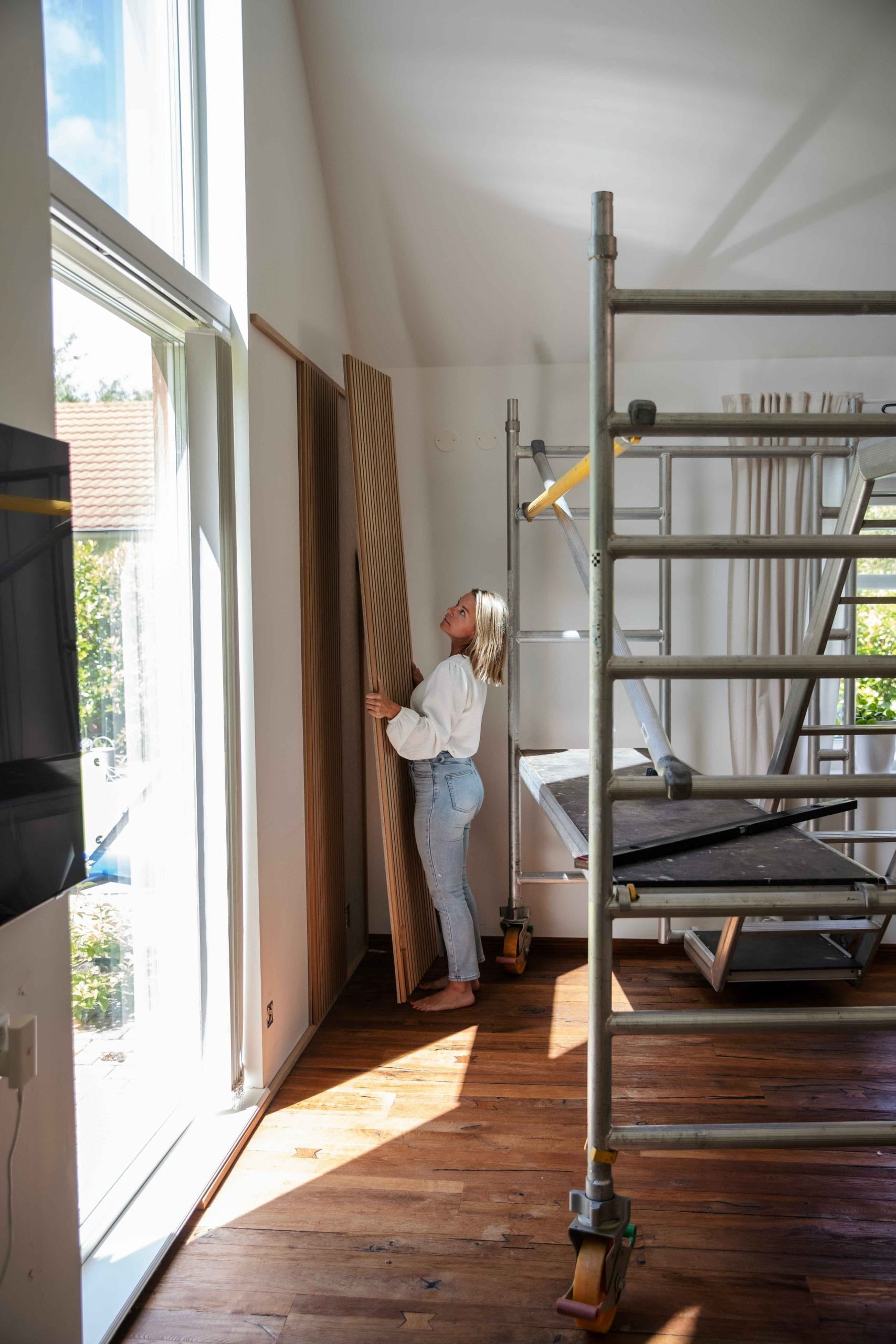 A woman stands in a sunlit room with large windows and scaffolding, holding an Akupanel acoustic wood panel on a polished wooden floor.