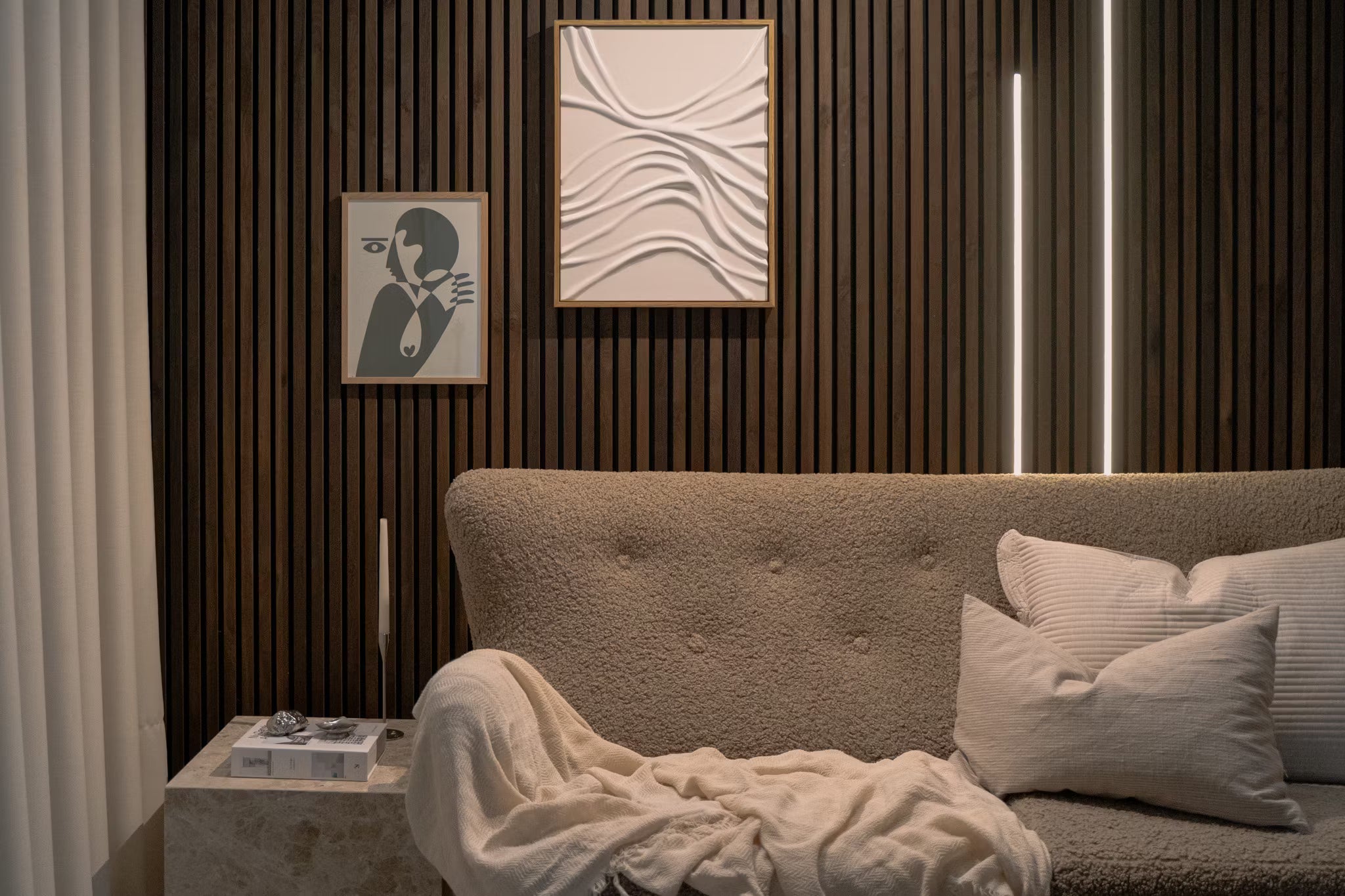 Modern living room with textured beige couch, white pillows, abstract acoustic panel art, and vertical Akupanel wood slat wall with soft ambient lighting.